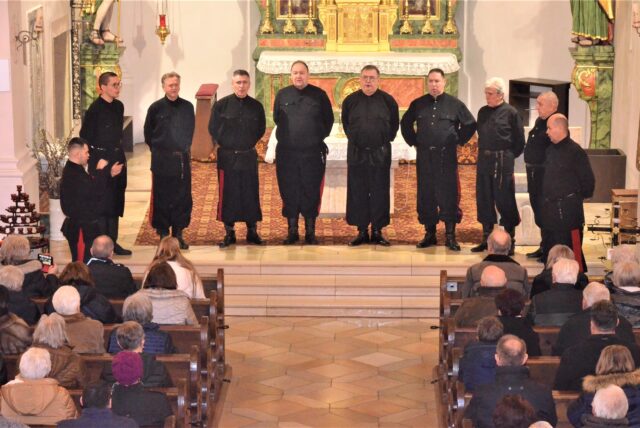 Eine Gruppe von Männern stehen in einer Kirche vor dem Altar und singen. Auf den Kirchenbänken sitzt das Publikum.