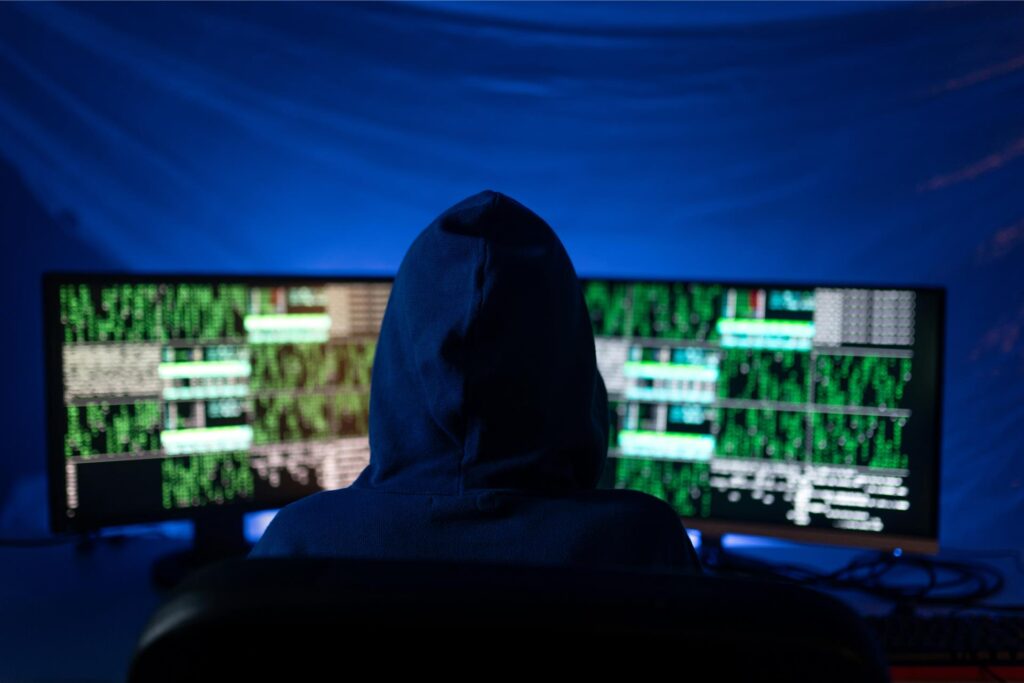 Ein Hacker im schwarzen Kapuzenpullover ist von hinten zu sehen und schaut auf zwei Bildschirme, auf denen Code zu sehen ist.