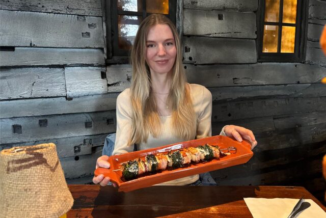 Eine Frau präsentiert einen Teller mit Spießen in einem Restaurant. Der Teller ist orange und die Spieße scheinen aus Fleisch und Gemüse zu bestehen.