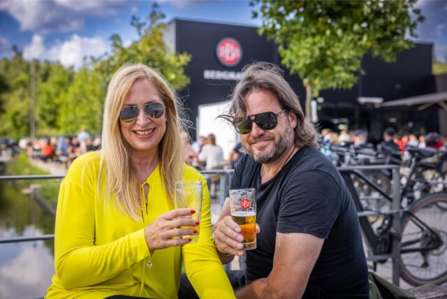 Zwei Personen sitzen im Freien und halten Gläser mit Bier. Im Hintergrund ist ein Gebäude mit dem Schriftzug „BERGMANN“ zu sehen.