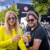 Zwei Personen sitzen im Freien und halten Gläser mit Bier. Im Hintergrund ist ein Gebäude mit dem Schriftzug „BERGMANN“ zu sehen.