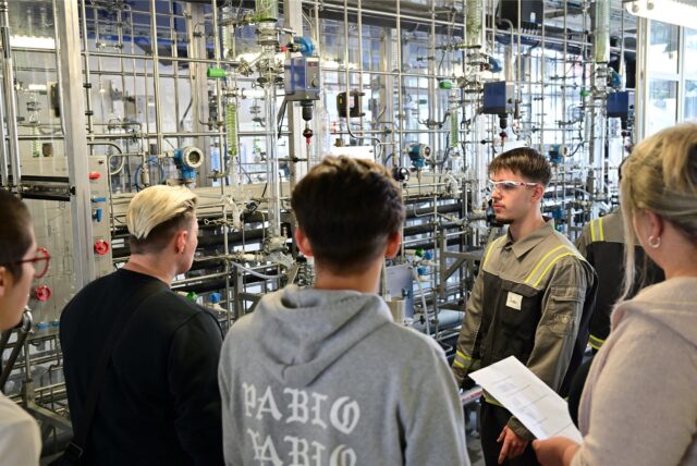 Mehrere Menschen stehen vor einer Anlage im Chemiepark. Ein Auszubildender in Sicherheitskleidung und mit Schutzbrille erklärt die Prozesse.