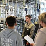 Mehrere Menschen stehen vor einer Anlage im Chemiepark. Ein Auszubildender in Sicherheitskleidung und mit Schutzbrille erklärt die Prozesse.