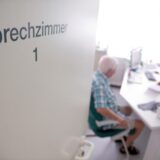 Blick in ein Sprechzimmer, in dem ein älterer Mann und eine Ärztin unscharf zu erkennen sind.