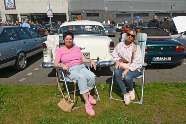 Zwei Frauen auf Campingstühlen vor Oldtimern