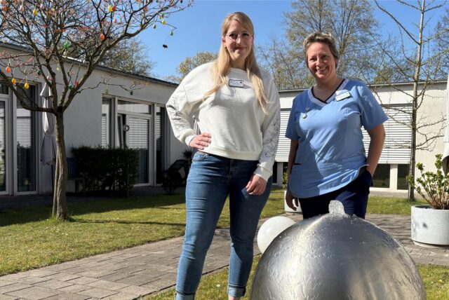 Andrea Nentwig und Denise Nonn stehen in einem Innenhof des Hospizes in Recklinghausen an einer großen, silbernen Kugel.