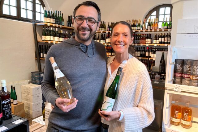 Sebastian Banhold und Daniela Münster stehen im Ladenlokal feinherb & konsorten und halten jeweils eine Flasche Wein in der Hand.