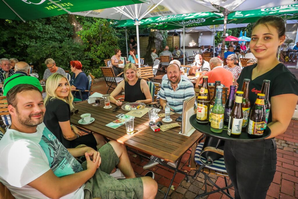 Eine Kellnerin steht lächelnd mit einem Tablet voller Getränke an einem Biergarten Tisch, an dem lachende Gäste sitzen. Eine Kellnerin steht lächelnd mit einem Tablet voller Getränke an einem Biergarten Tisch, an dem lachende Gäste sitzen.