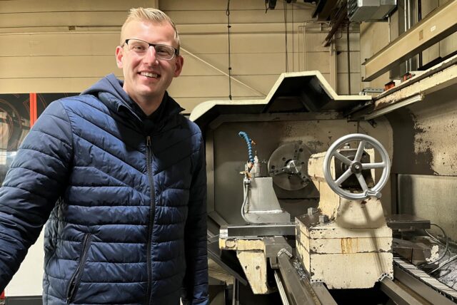 Ein junger Mann mit blonden Haaren steht an einer Drehmaschine und lächelt in die Kamera.