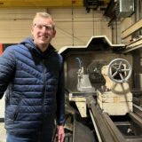 Ein junger Mann mit blonden Haaren steht an einer Drehmaschine und lächelt in die Kamera.