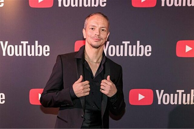 DJ Alle Farben im schwarzen Anzug vor einer Wand mit YouTube-Logos darauf