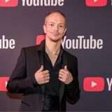 DJ Alle Farben im schwarzen Anzug vor einer Wand mit YouTube-Logos darauf