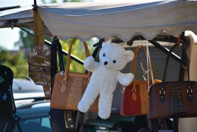 Auf dem Bild sind verschiedene Artikel, darunter Handtaschen, ein Rucksack und ein Teddybär, an einem Verkaufsstand aufgehängt. Die Artikel werden mit Klemmen an der Überdachung des Standes befestigt.