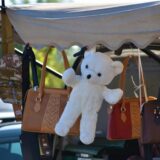 Auf dem Bild sind verschiedene Artikel, darunter Handtaschen, ein Rucksack und ein Teddybär, an einem Verkaufsstand aufgehängt. Die Artikel werden mit Klemmen an der Überdachung des Standes befestigt.