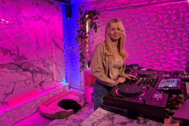Eine junge Frau steht an einem DJ-Pult in einem beleuchteten Raum mit verschiedenen Dekorationen und Graffiti an den Wänden. Im Vordergrund befindet sich ein Hundebett mit einem schlafenden schwarzen Hund.
