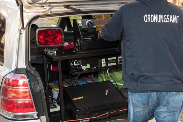 Zu sehen ist ein Auto mit geöffneten Kofferraum, in dem sich eine mobile Verkehrsüberwachung befindet. Rechts im Bild steht eine Person, die eine Jacke mit der Aufschrift „Ordnungsamt“ trägt.