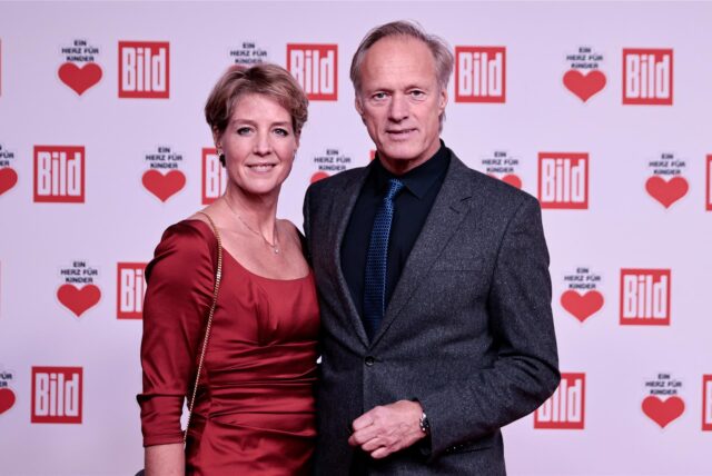 Gerhard Delling und Christina Block stehen vor einer Werbewand bei der Spendengala „Ein Herz für Kinder“ auf dem Roten Teppich an.