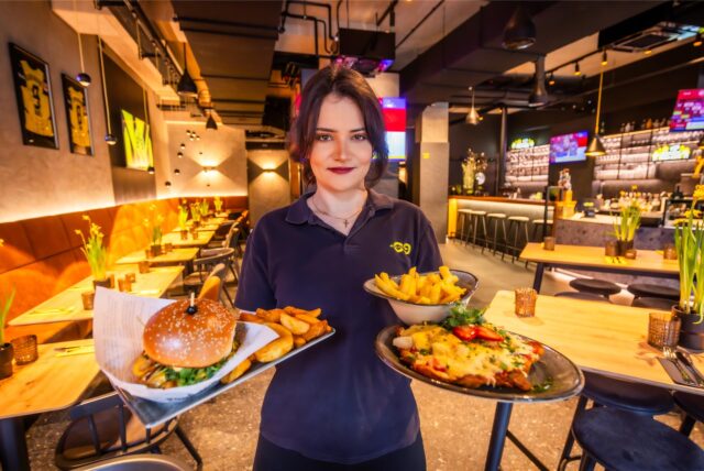 Eine Mitarbeiterin des Dortmunder Bistro 09 hält Teller mit Burger, Pommes und Schnitzel lächelnd in die Kamera