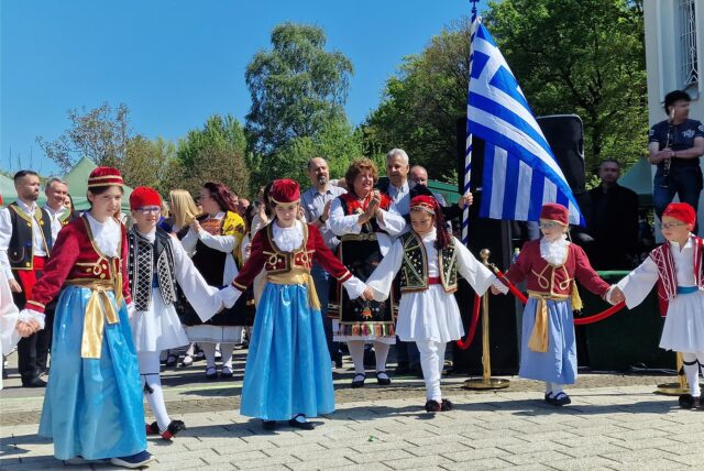 Kinder in Trachtenkleidung führen einen Tanz auf. Dahinter ist die griechische Nationalflagge zu sehen.