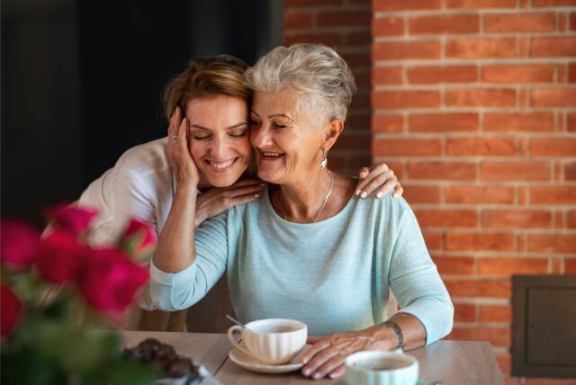 Eine Frau umarmt ihre Mutter von hinten zum Muttertag. Auf dem Tisch vor ihnen stehen Kaffeetassen und Blumen.