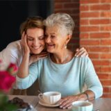 Eine Frau umarmt ihre Mutter von hinten zum Muttertag. Auf dem Tisch vor ihnen stehen Kaffeetassen und Blumen.
