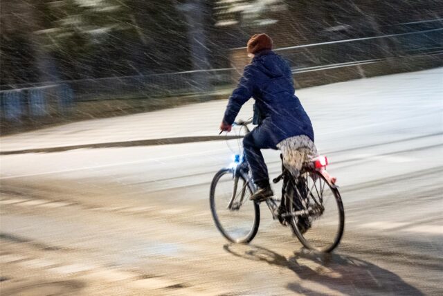 Eine Fahrradfahrerin fährt am Abend bei Schneefall über eine Straße