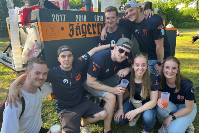 Eine Gruppe junger Menschen sitzt, steht vor dem Jägermeisterwagen und grinsen in die Kamera.