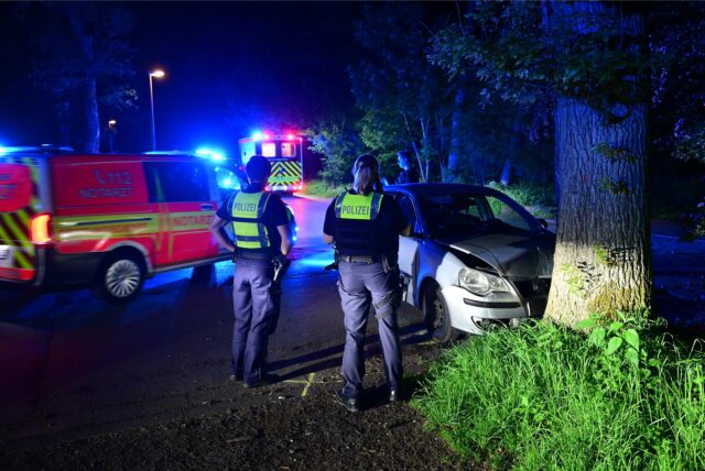 Polizisten stehen vor einem Auto, das gegen einen Baum geprallt ist.