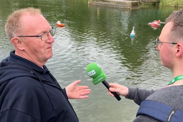 Zwei Männer stehen auf einem Steg und sprechen miteinander. Im Hintergrund sind Modellboote auf dem Wasser zu sehen.