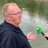 Zwei Männer stehen auf einem Steg und sprechen miteinander. Im Hintergrund sind Modellboote auf dem Wasser zu sehen.
