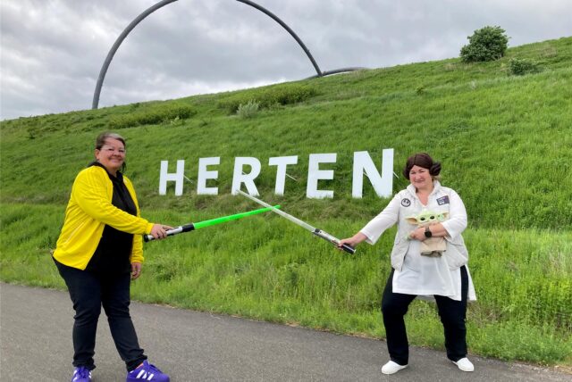 Heidi Fischer (r.) und ihre Nichte Sonja Kipper halten Laser-Schwerter und eine Stars-Wars-Figur in den Händen und posieren vor dem Herten-Schriftzug auf der Halde Hoheward in Herten.