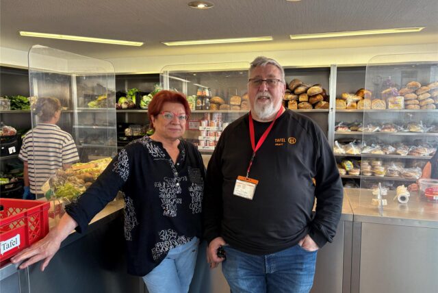 Edeltraud Hartelt und Wolfgang Schulz stehen im Vordergrund. Hinter ihnen sind die gefüllten Regale der Tafel zu sehen.