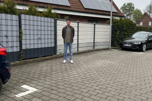 Mert Erdem steht auf einer leeren Fläche zwischen zwei belegten Parkplätzen am Fahrbahnrand.