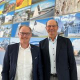 Zwei Männer im Anzug stehen nebeneinander vor einer Wand mit verschiedenen Bildern.