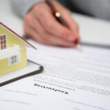 Ein Mann unterschreibt den Kaufvertrag für ein Einfamilienhaus, dessen Modell links daneben steht.
