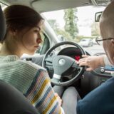 Eine junge Frau sitzt am Steuer eines VW und wir von einem Fahrlehrer instruiert.