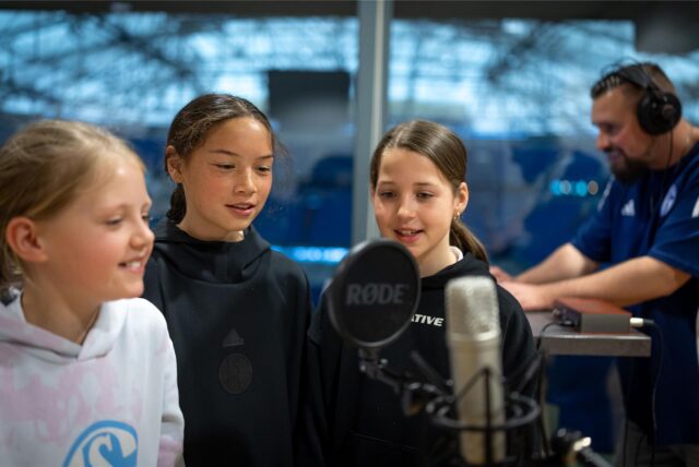 Drei Mädchen stehen in der Nordkurve der Veltins-Arena vor einem Mikrofon, im Hintergrund steht Seba mit Kopfhörern an einem Rechner.