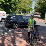 Ein Mann mit Leuchtweste steht an seinem Fahrrad. Im Hintergrund biegen Autos auf einem Parkplatz ab.