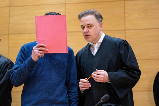 Anwalt Burkhard Benecken mit einem Angeklagten, der sich einen Ordner vor das Gesicht hält.