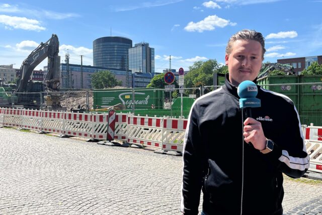 Tim Hübbertz steht mit einem Mikrofon in der Hand in der Dortmunder Nordstadt und berichtet über eine Großbaustelle.