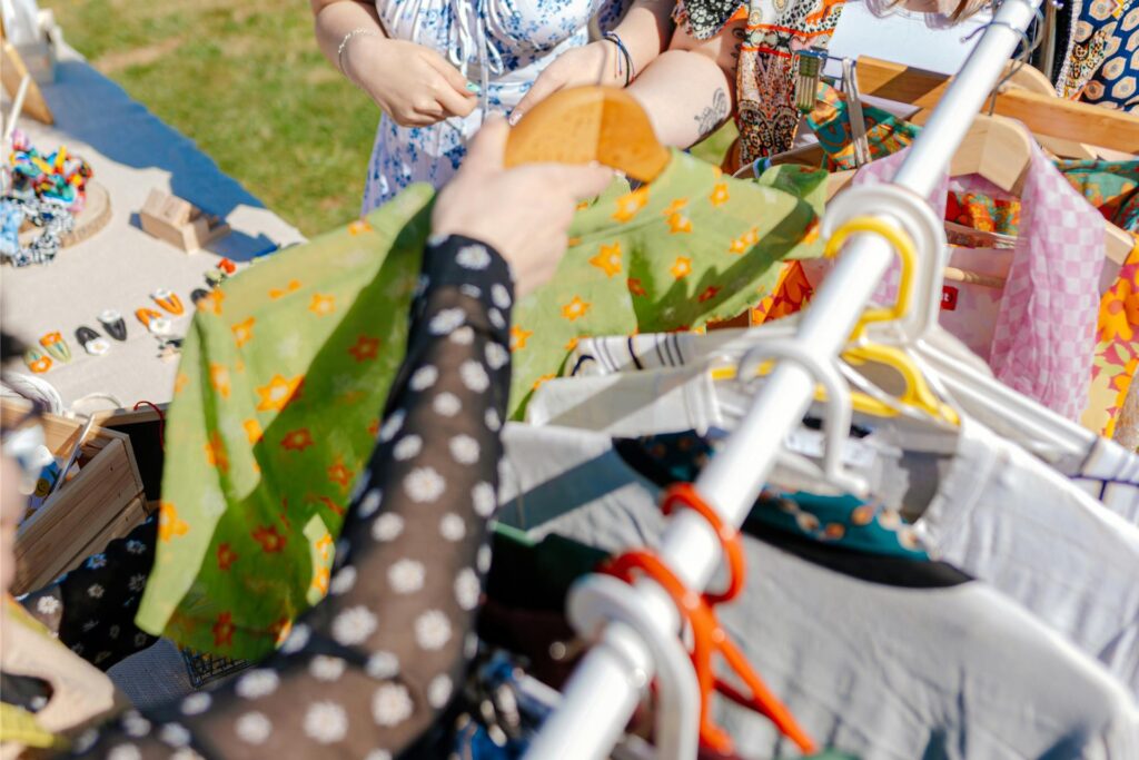Auf einem Trödelmarkt-Stand liegen verschiedenste Accessoires - daneben steht eine Kleiderstange mit bunten Blusen, T-Shirts und weiteren gebrauchten Kleidungsstücken.