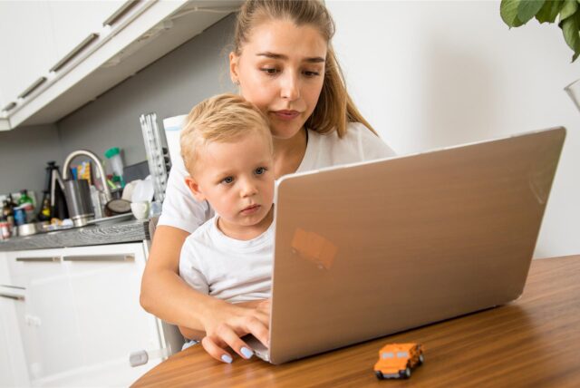 Eine junge Frau sitzt mit ihrem Laptop an einem Tisch und hat ihren Sohn auf dem Schoß.