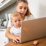 Eine junge Frau sitzt mit ihrem Laptop an einem Tisch und hat ihren Sohn auf dem Schoß.