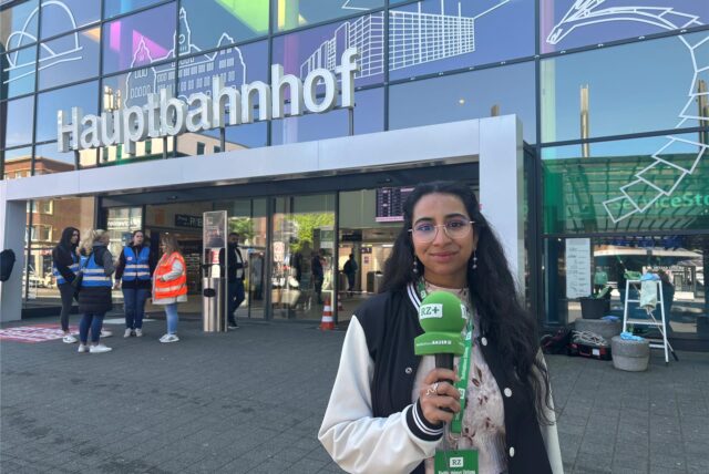 Die Front des Hauptbahnhofs Recklinghausen. Davor steht Reporterin Rosi Taslima Azam mit einem Mikrofon in der Hand.