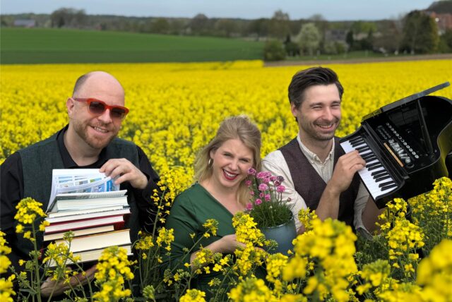 Michael Baute hält einen Stapel Bücher in den Händen, Tamara Peters hält einen Blumentopf und Florian Albers hält ein Mini-Piano. Zusammen stehen sie in einem Rapsfeld.