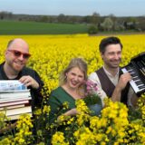 Michael Baute hält einen Stapel Bücher in den Händen, Tamara Peters hält einen Blumentopf und Florian Albers hält ein Mini-Piano. Zusammen stehen sie in einem Rapsfeld.