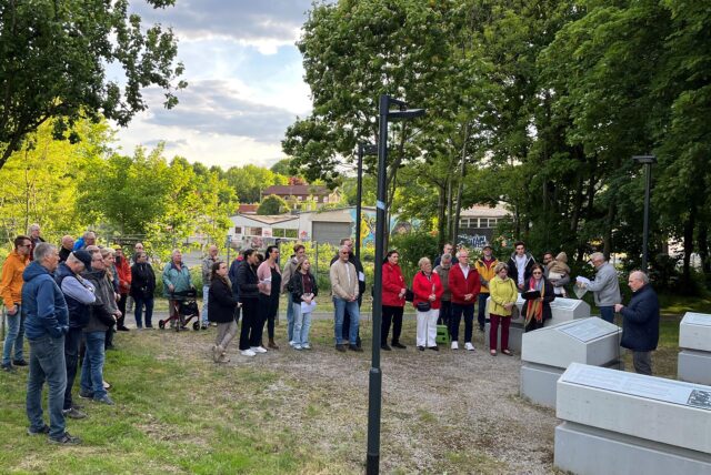Eine Gruppe von Menschen versammelt sich im Freien, um einer Rede zuzuhören. Im Vordergrund sind Betonblöcke mit angebrachten Informationstafeln zu sehen.