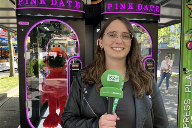 Eine junge Frau mit Brille hält ein Mikrofon des Medienhauses Bauer in der Hand. Im Hintergrund befinden sich zwei Spielautomaten mit der Aufschrift „Pink Date“.