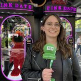 Eine junge Frau mit Brille hält ein Mikrofon des Medienhauses Bauer in der Hand. Im Hintergrund befinden sich zwei Spielautomaten mit der Aufschrift „Pink Date“.