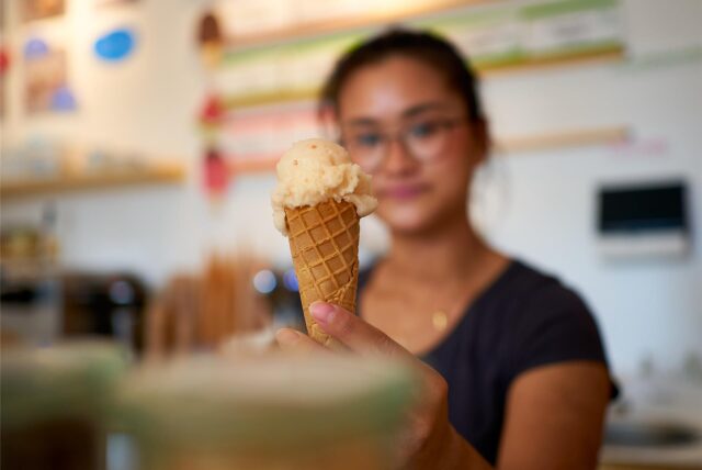 Eine junge Frau hält eine Eistüte mit zwei Kugeln Eis. Das Eis ist hellbeige und die Waffel hat ein Wabenmuster.